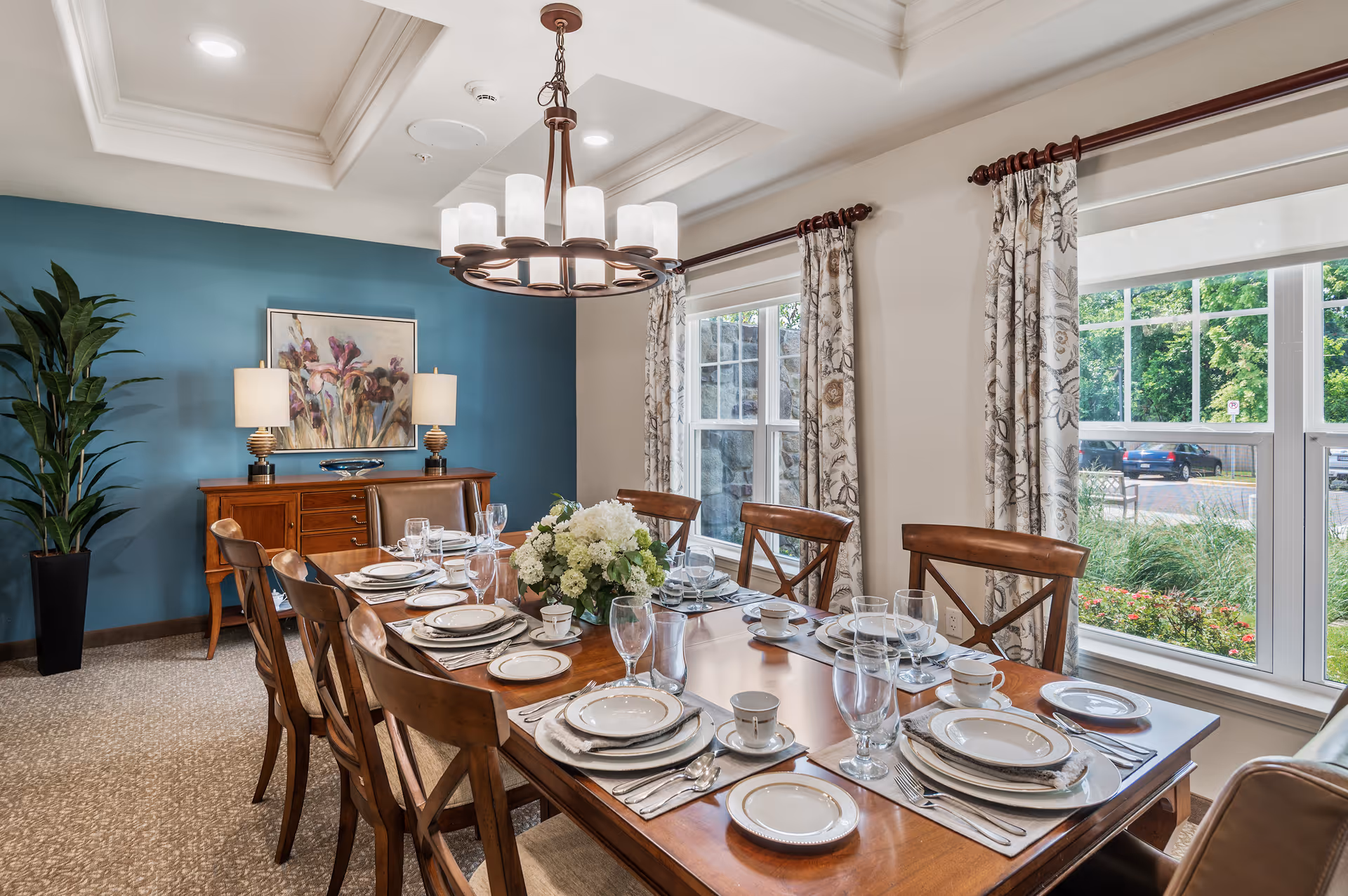 A well-lit dining room with a wooden dining table set for eight people with plates, cups, glasses, and silverware. The room features a blue accent wall with a painting and two table lamps on a wooden sideboard. Large windows with floral curtains allow natural light to brighten the space, and a decorative chandelier hangs from the ceiling.