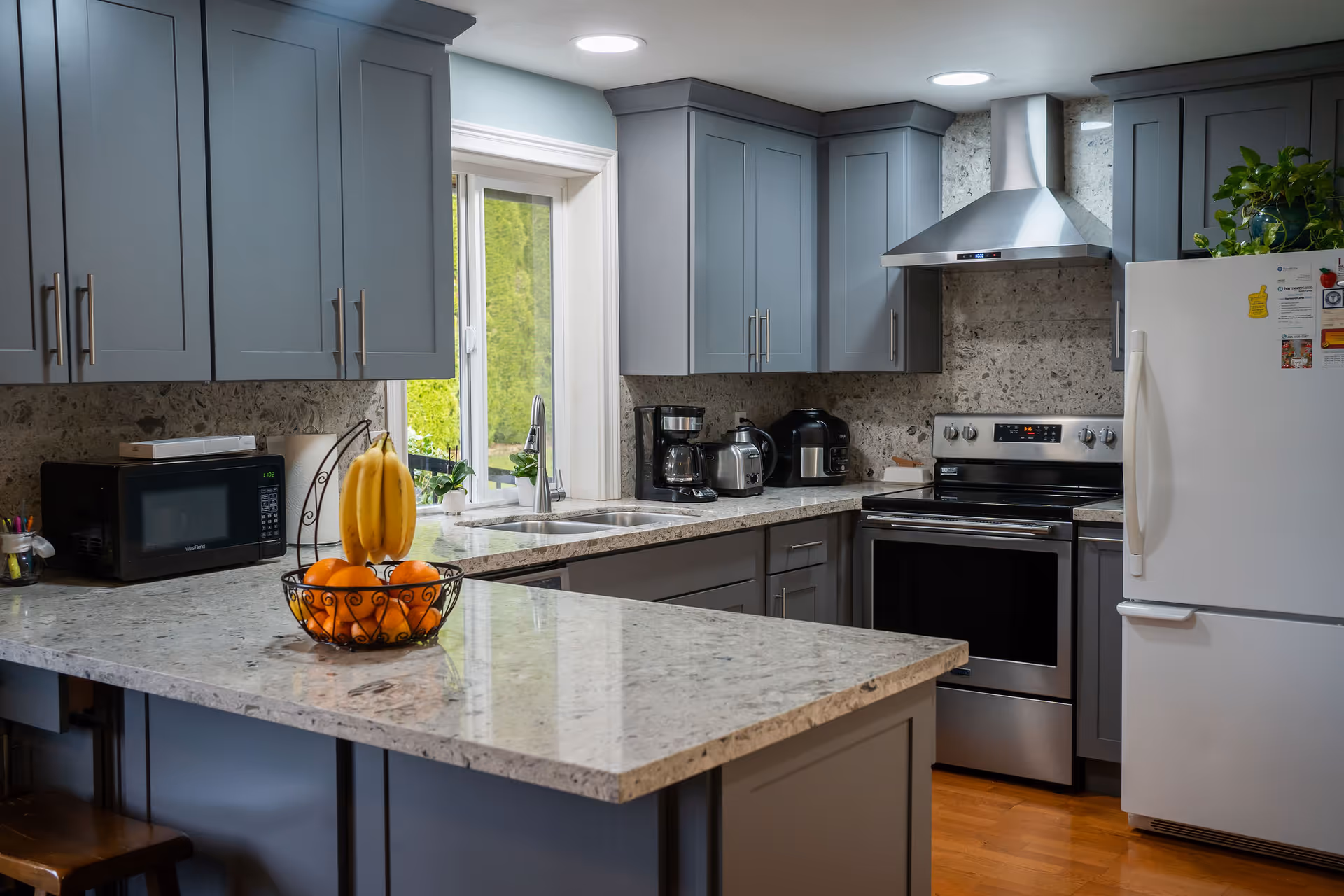 Modern kitchen with gray cabinets, marble countertops, stainless steel stove and hood, white refrigerator, and a fruit bowl on the island.