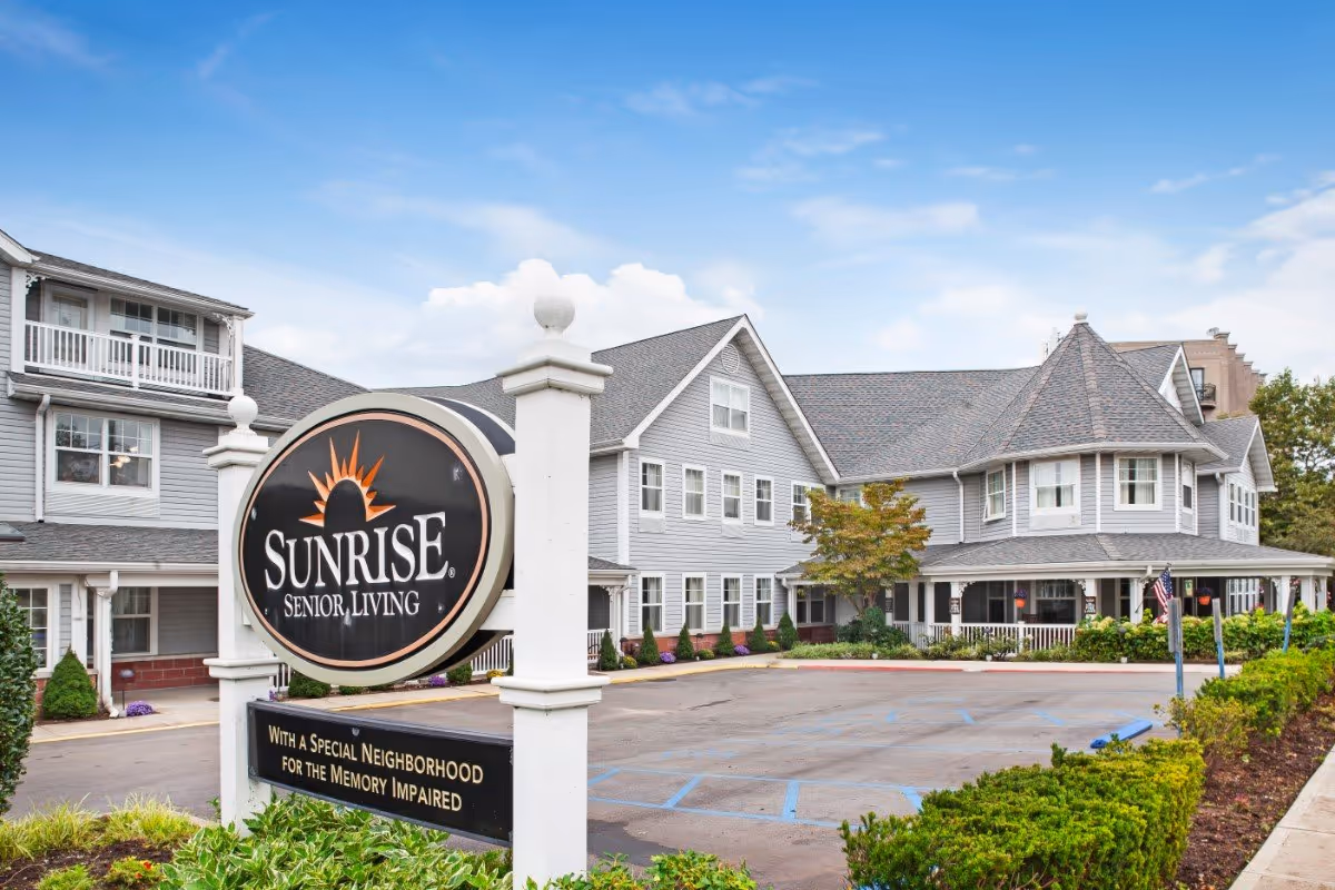 Front exterior of the Sunrise Senior Living building with a large round sign, driveway, and landscaped entrance.