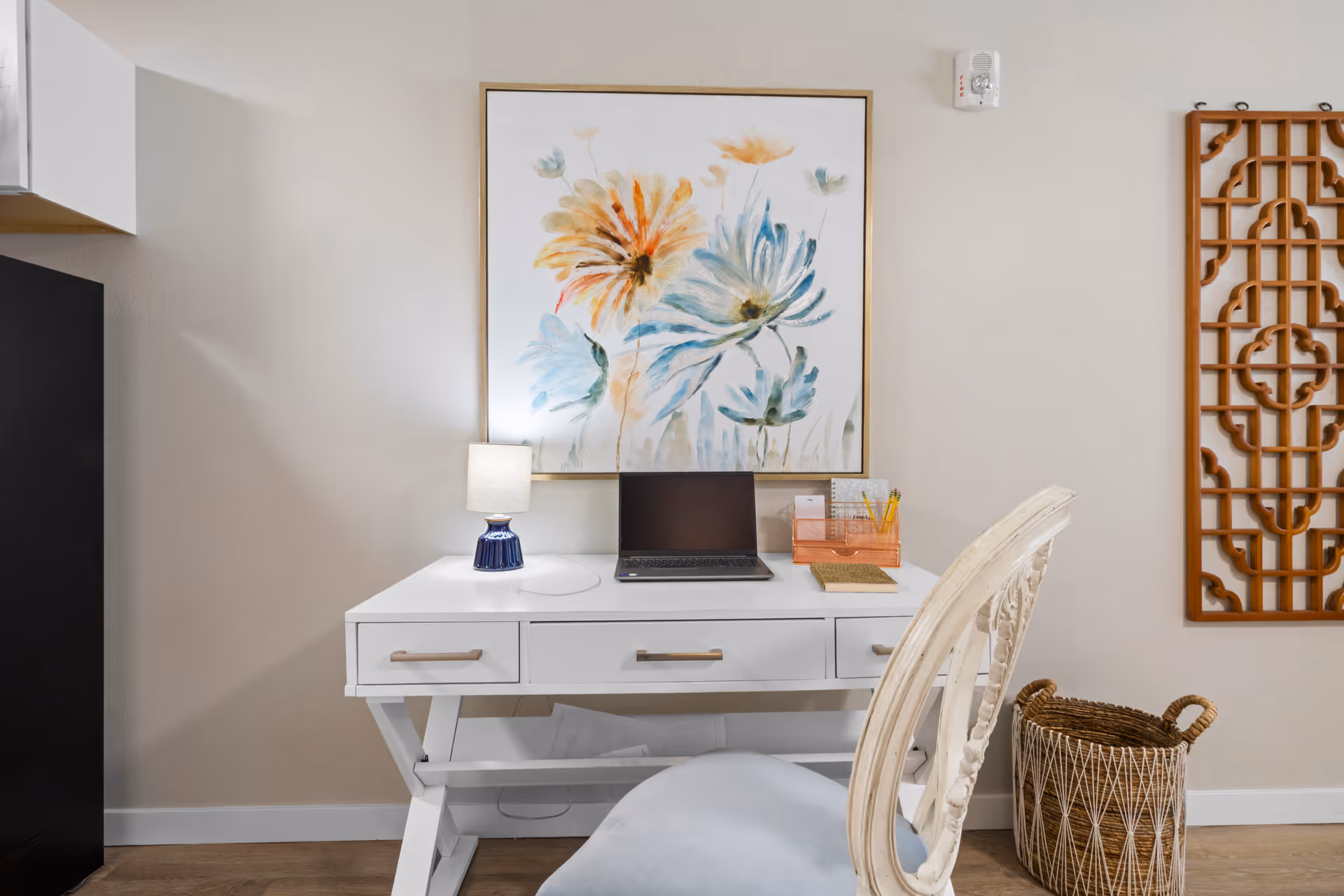 A white desk with three drawers featuring a small lamp, a closed laptop, and an organizer with pencils and papers. Above the desk hangs a large floral painting with blue and yellow flowers. To the right of the desk is a decorative wooden wall piece and a woven basket on the floor. A white chair with a cushioned seat is positioned in front of the desk.