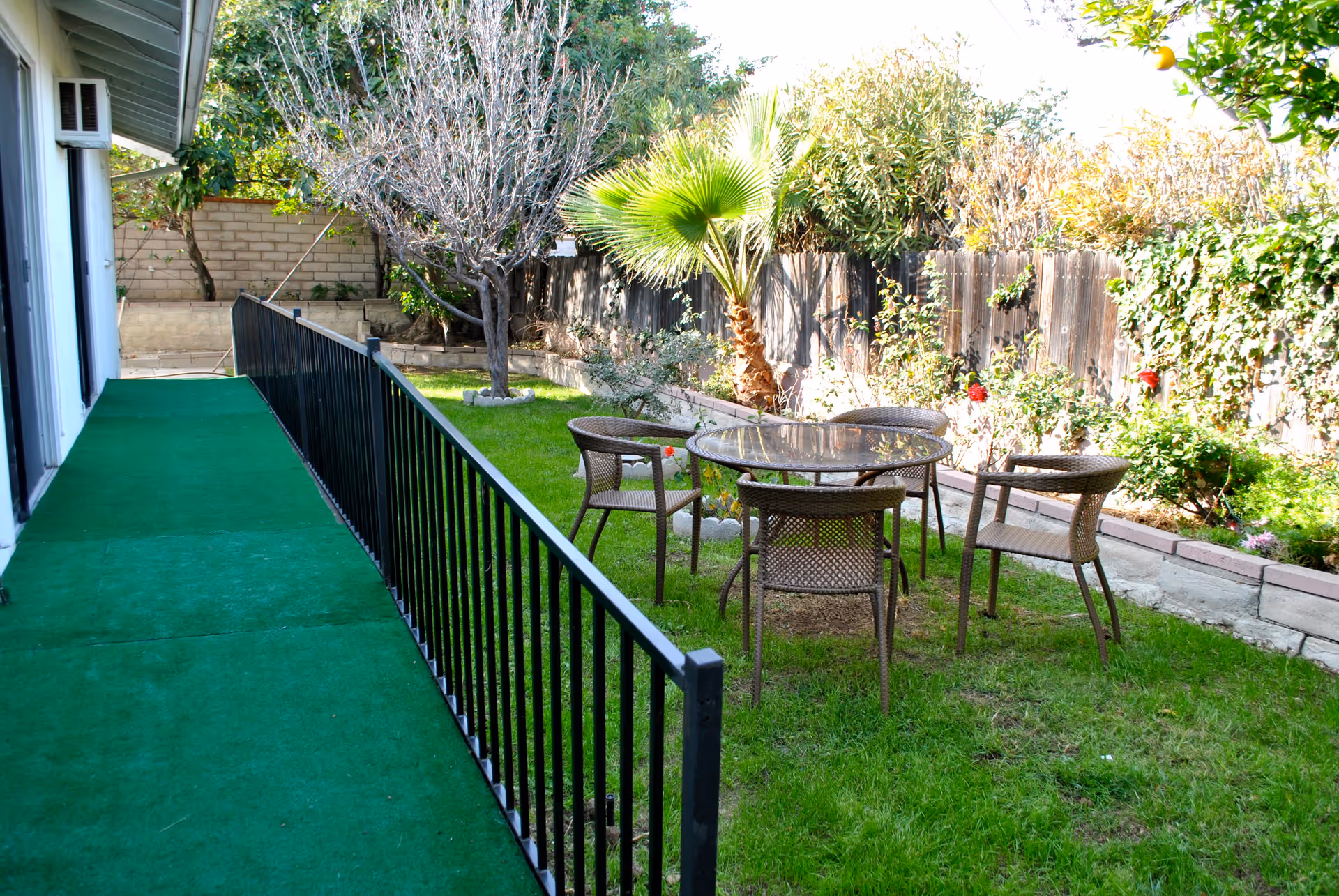 Outdoor garden area with a green artificial turf pathway alongside a building. There is a black metal railing separating the pathway from a grassy lawn with a round glass table and four wicker chairs. The garden is bordered by a wooden fence with various plants and trees, including a palm tree and a leafless tree.