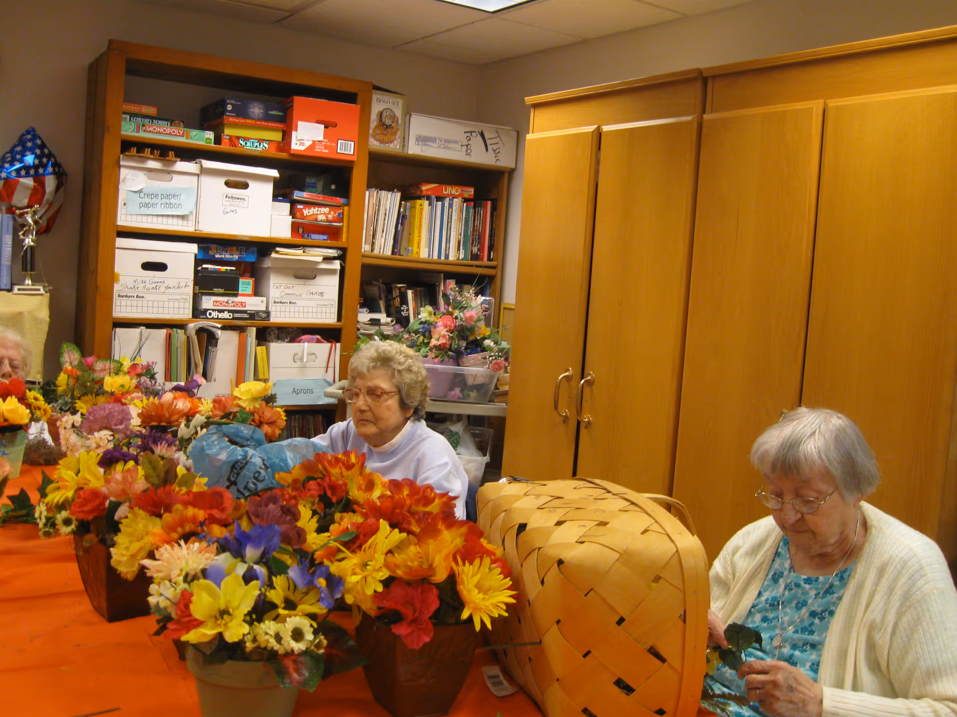 Two elderly women sitting at a table covered with colorful artificial flowers in a room with wooden cabinets and shelves filled with board games, books, and storage boxes.