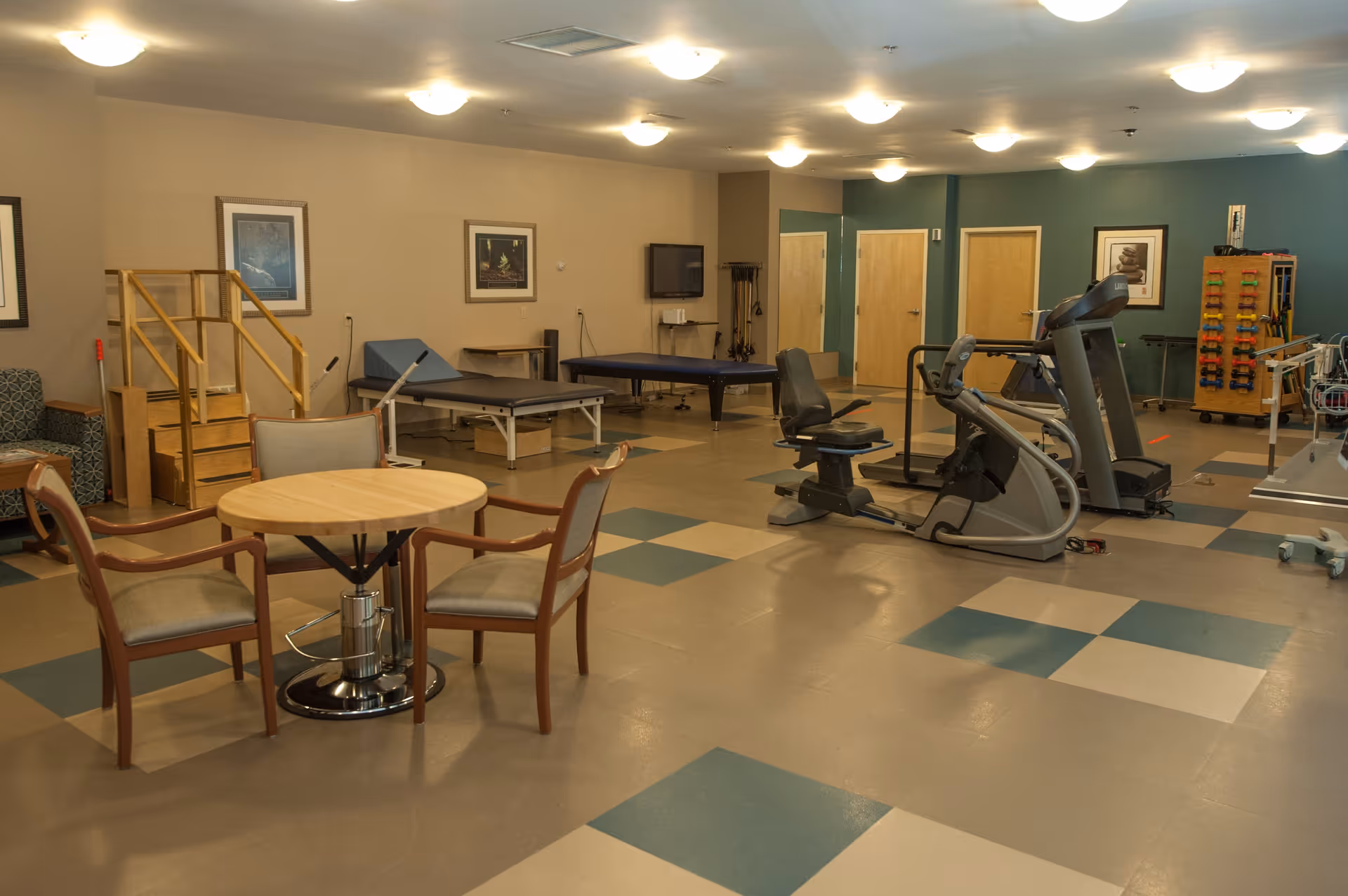 A spacious therapy or exercise room with various equipment including a recumbent exercise bike, treadmill, therapy tables, and a small set of stairs for physical therapy. There is a round table with four chairs in the foreground, framed pictures on the walls, and a rack with colorful hand weights in the background.