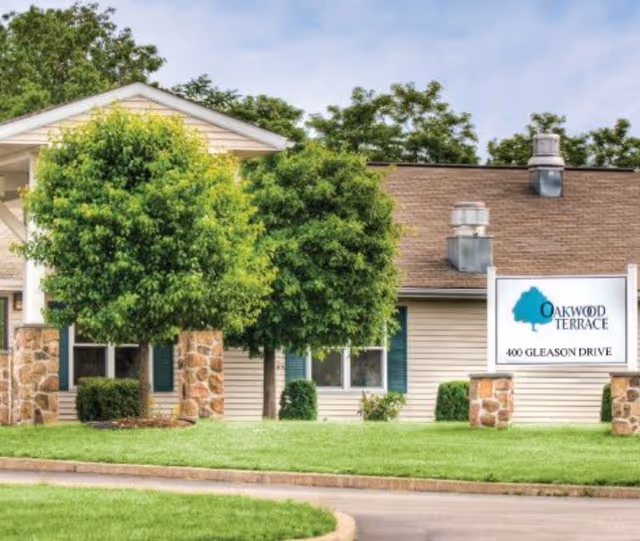 Front exterior of the Oakwood Terrace building with trees, lawn, and a sign reading 'Oakwood Terrace 400 Gleason Drive'.