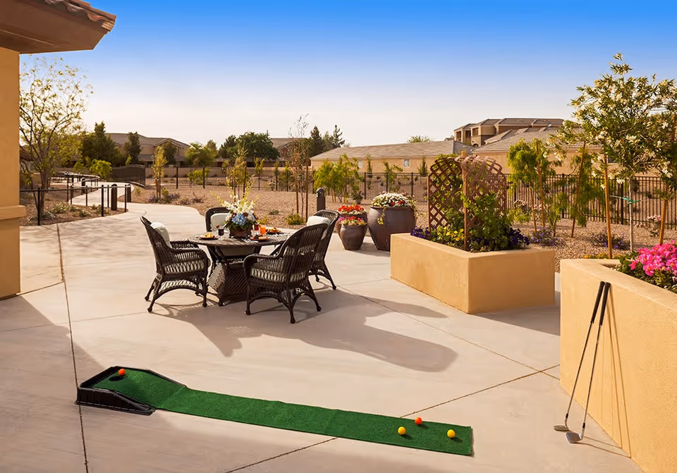 Outdoor patio area with a round table and four wicker chairs, decorated with a flower arrangement and refreshments. There are potted plants and raised garden beds with flowers and greenery. A small putting green with golf balls and clubs is set up on the concrete patio. The background shows a fenced area with trees and buildings under a clear blue sky.