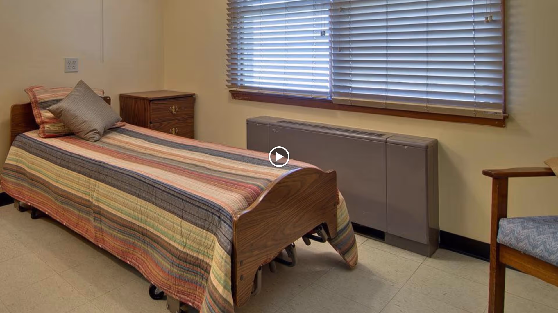A simple bedroom with a single wooden bed covered in a striped quilt, a bedside table, a chair, and a window with blinds.
