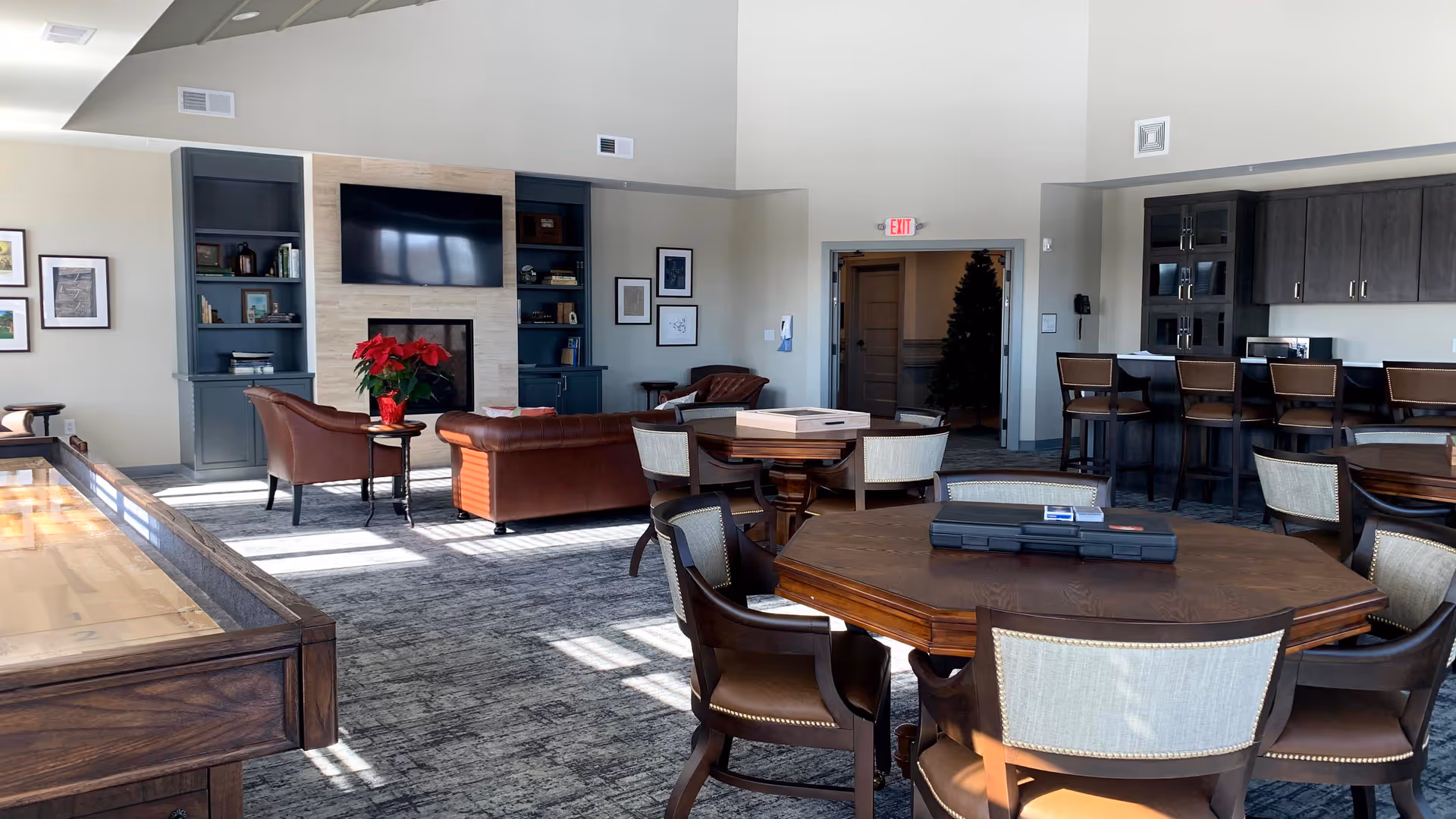 A spacious common area with a shuffleboard table on the left, several round wooden tables with chairs, a seating area with brown leather sofas and armchairs in front of a wall-mounted TV and fireplace, built-in shelves with books and decor, and a kitchen counter with bar stools on the right. The room has high ceilings and large windows allowing natural light to fill the space.