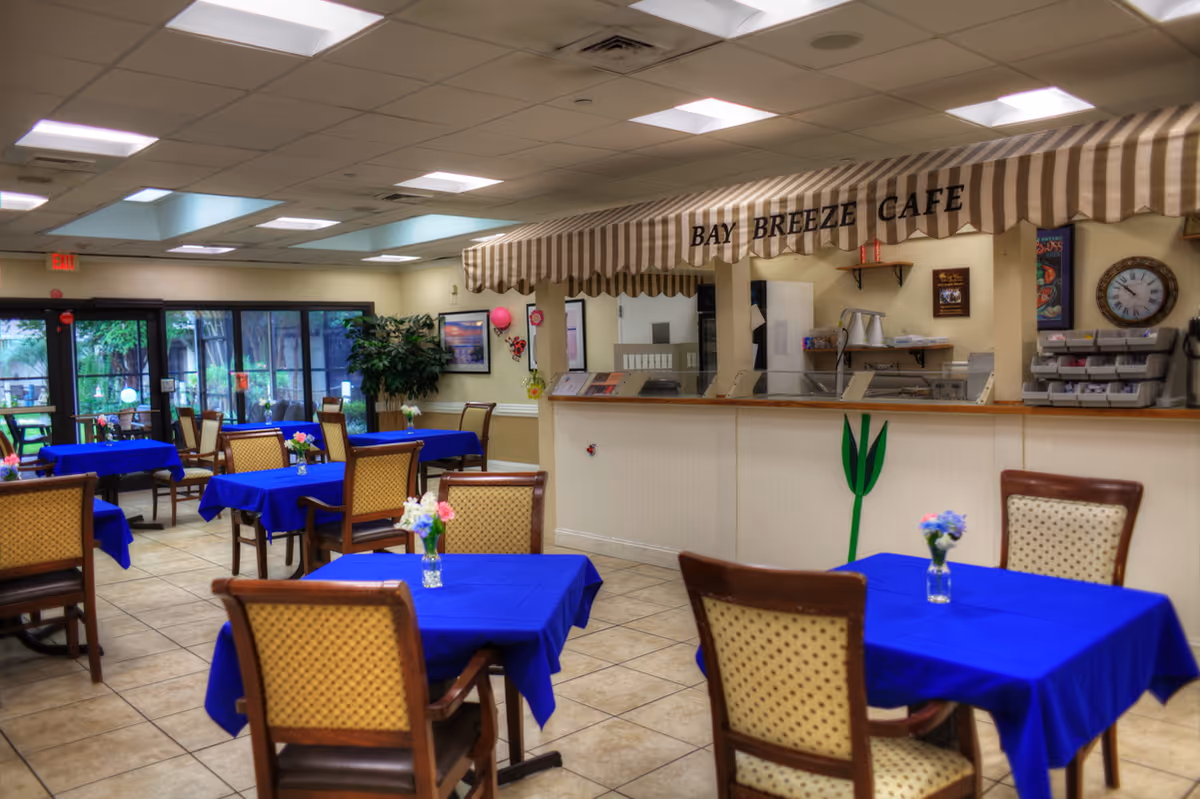 Interior view of Bay Breeze Cafe in a senior living facility, showing multiple tables covered with blue tablecloths and small flower vases. There are wooden chairs with cushioned seats around the tables. The cafe counter has a striped beige and white awning with the name 'Bay Breeze Cafe' displayed. The room has tiled flooring, ceiling lights, and large windows with a view of greenery outside.
