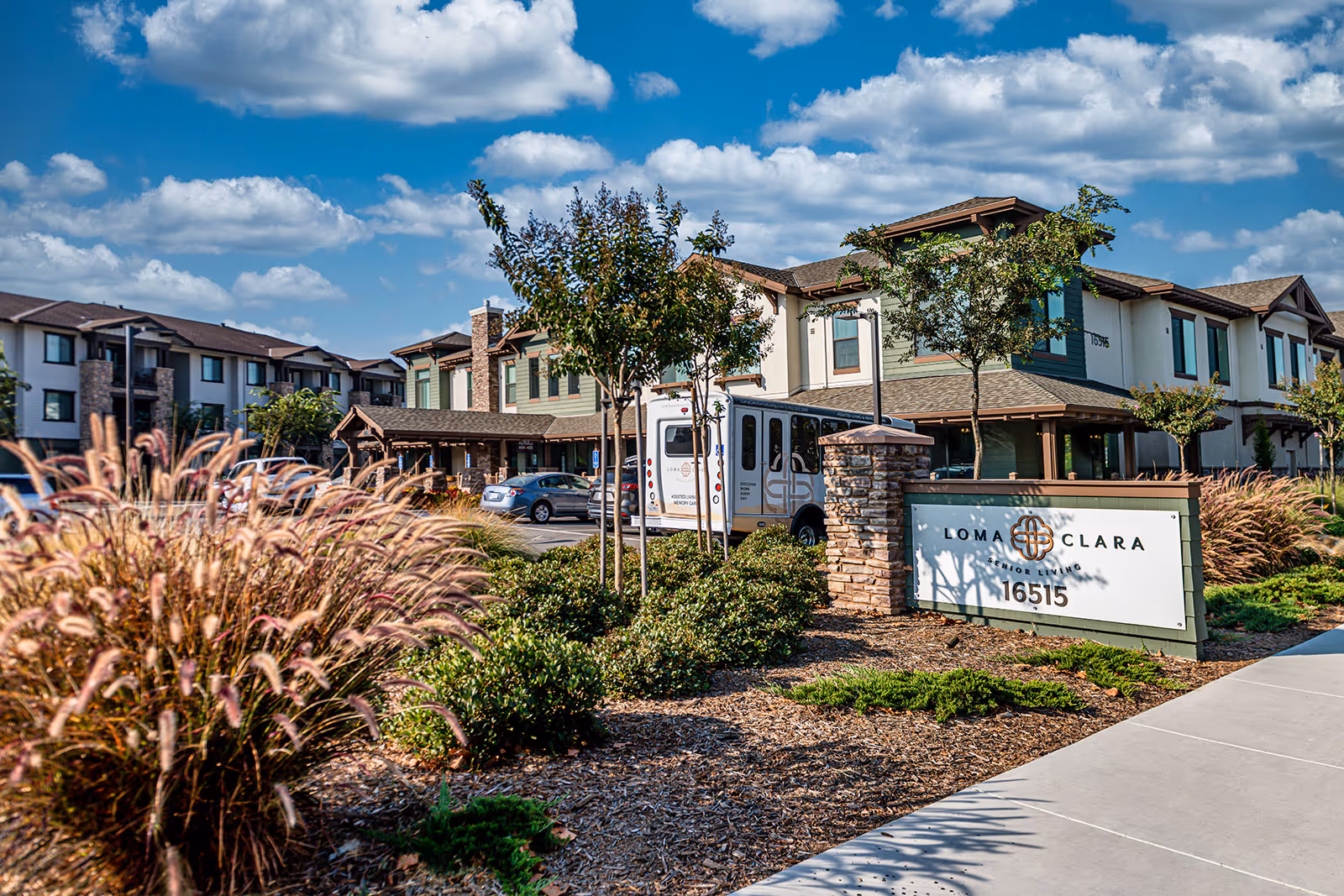 Exterior view of Loma Clara Senior Living facility showing a two-story building with green and beige siding, a parking lot with cars and a shuttle bus, landscaped bushes and ornamental grasses, and a clear blue sky with scattered clouds.