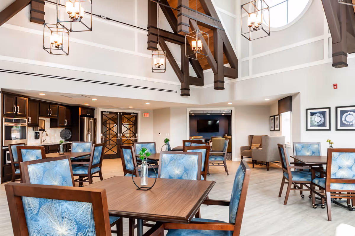 A bright and spacious dining area in Arbor Terrace Mount Laurel featuring wooden tables with blue patterned chairs. The room has high ceilings with exposed dark wooden beams and modern hanging light fixtures. There is a kitchen area with dark cabinetry and stainless steel appliances on the left side, and a seating area with armchairs and framed artwork on the right. Large windows allow natural light to fill the space.