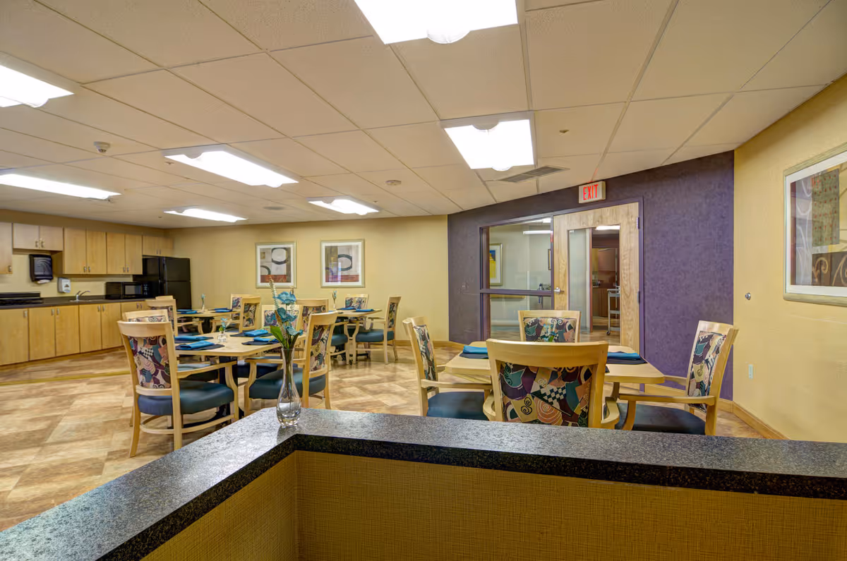 Communal dining room with round tables set with teal napkins, patterned wooden chairs, a kitchenette on the left, and a purple accent wall with an exit door.