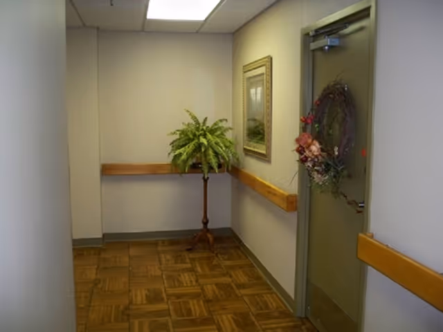 A small hallway corner in a senior living facility with wooden handrails on the walls, a green door decorated with a floral wreath, a framed picture on the wall, and a potted fern plant on a wooden stand.
