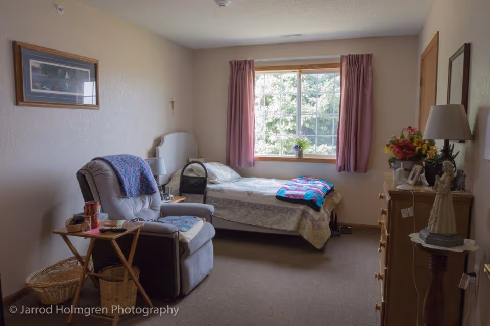 A cozy senior bedroom with a single bed, recliner, dresser, and a window with pink curtains.
