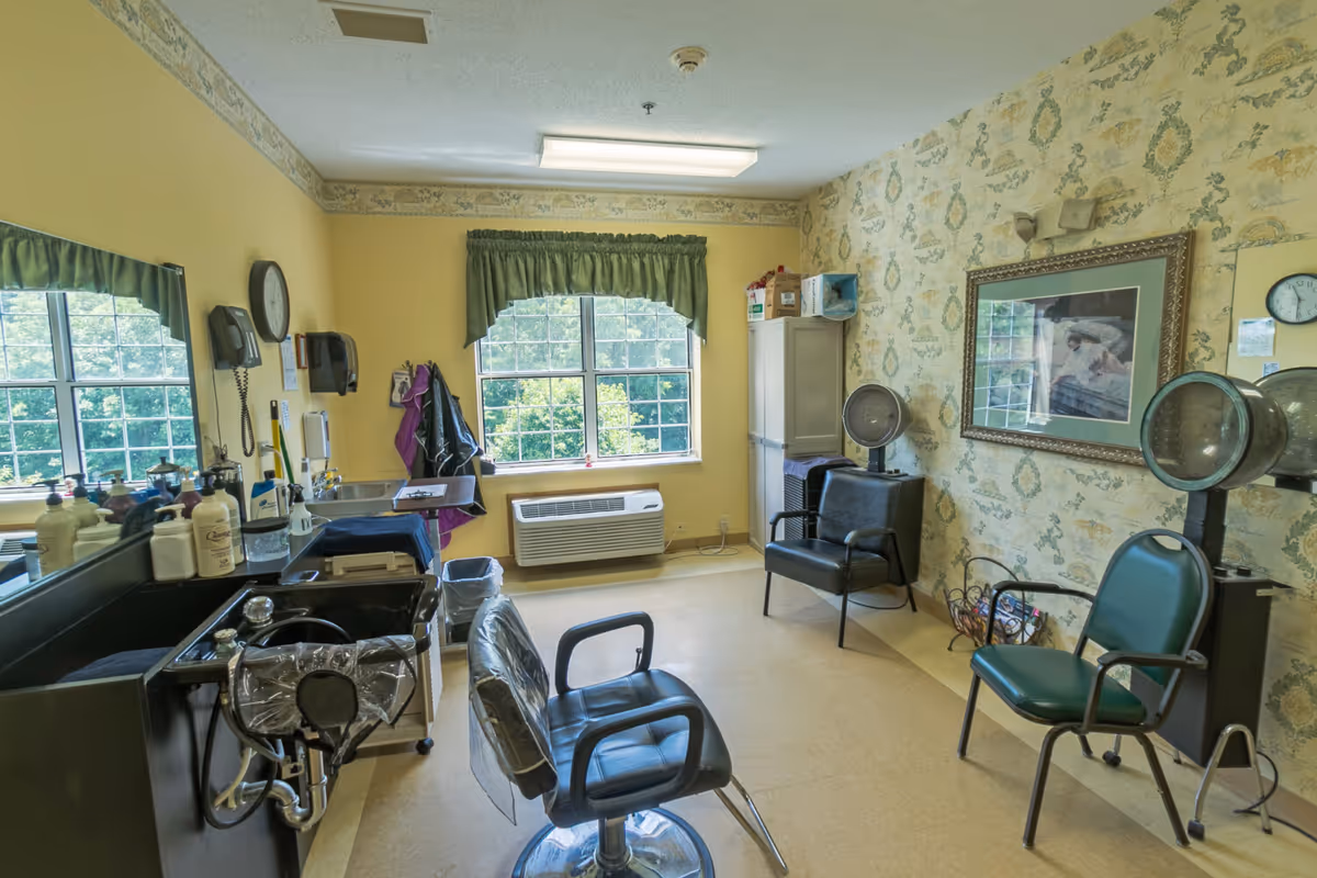 A small salon room with two black salon chairs, two hair drying machines, a black sink with hair washing equipment, a wall clock, a window with green curtains, and a framed picture on the wall with floral wallpaper.