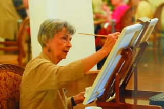 An elderly woman painting on a canvas set on an easel in a warmly lit indoor setting, with blurred people and furniture in the background.
