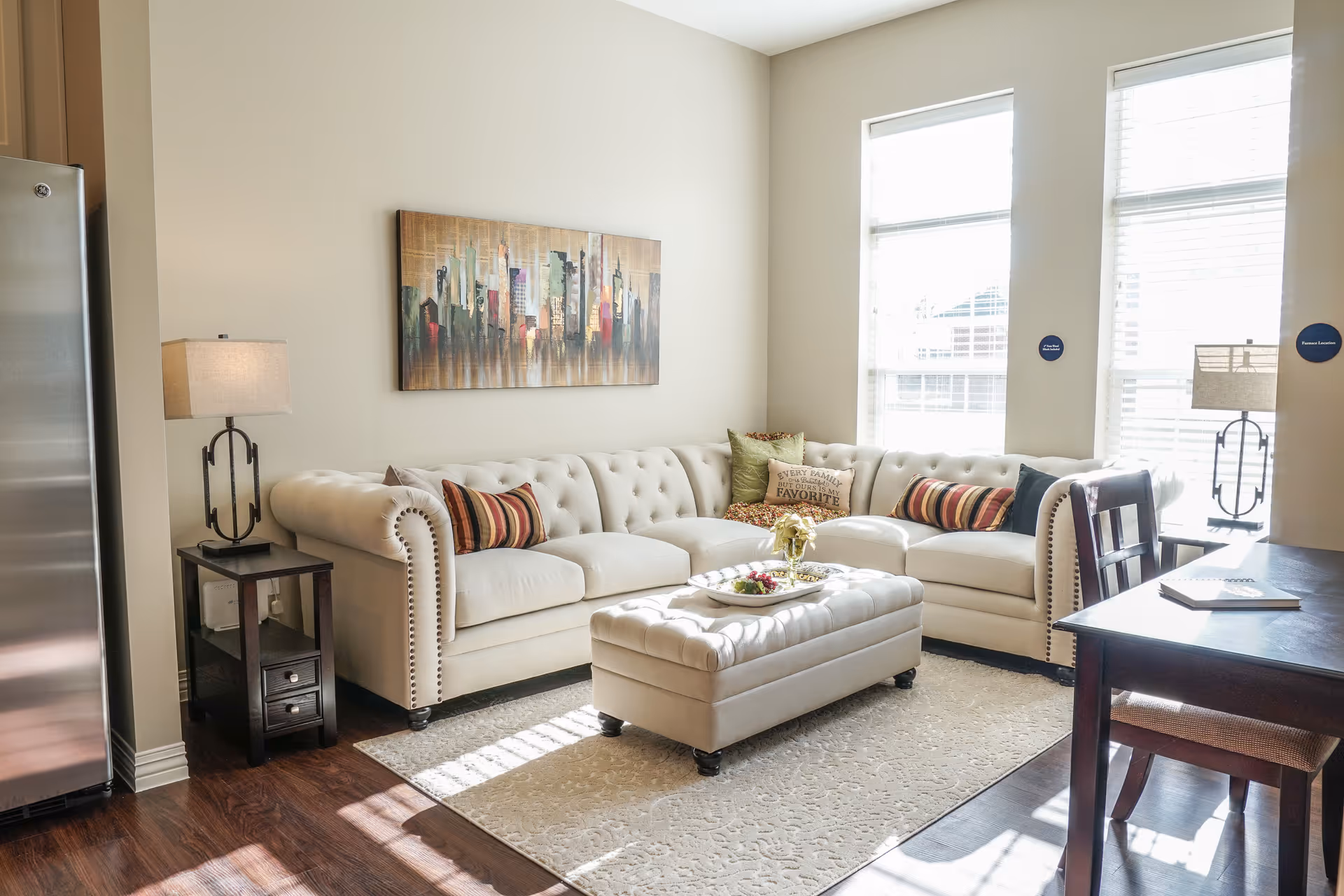 A bright living room with a large cream-colored tufted sectional sofa adorned with striped and patterned throw pillows. A matching tufted ottoman sits in front of the sofa on a light-colored rug. There is a dark wooden side table with a lamp next to the sofa and a dark wooden dining table with chairs on the right. Two large windows with blinds allow natural light to fill the room. A colorful cityscape painting hangs on the wall above the sofa.
