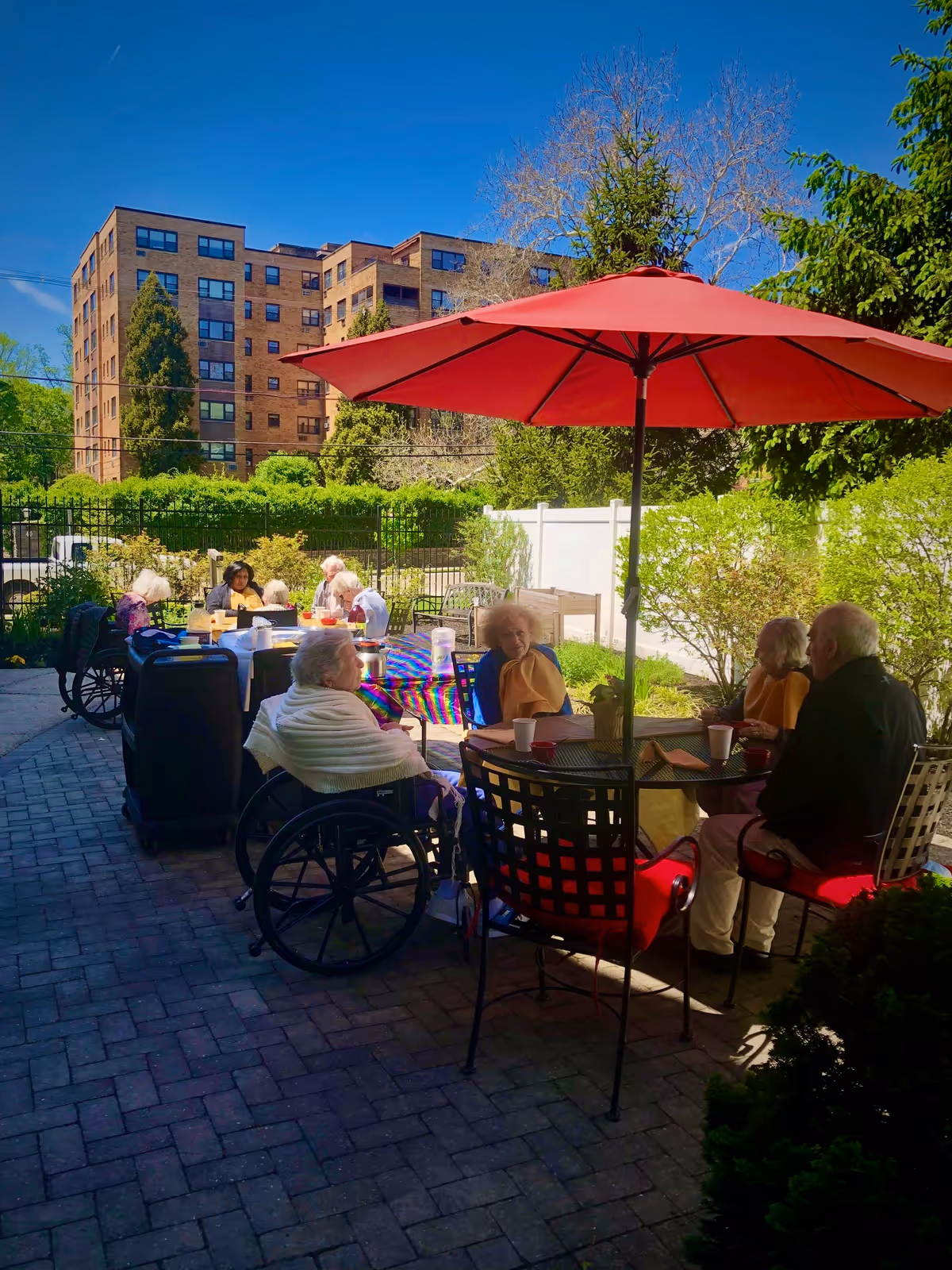 Older adults sit at patio tables under a red umbrella in a sunny courtyard outside a senior living facility with a multi-story brick building behind them.