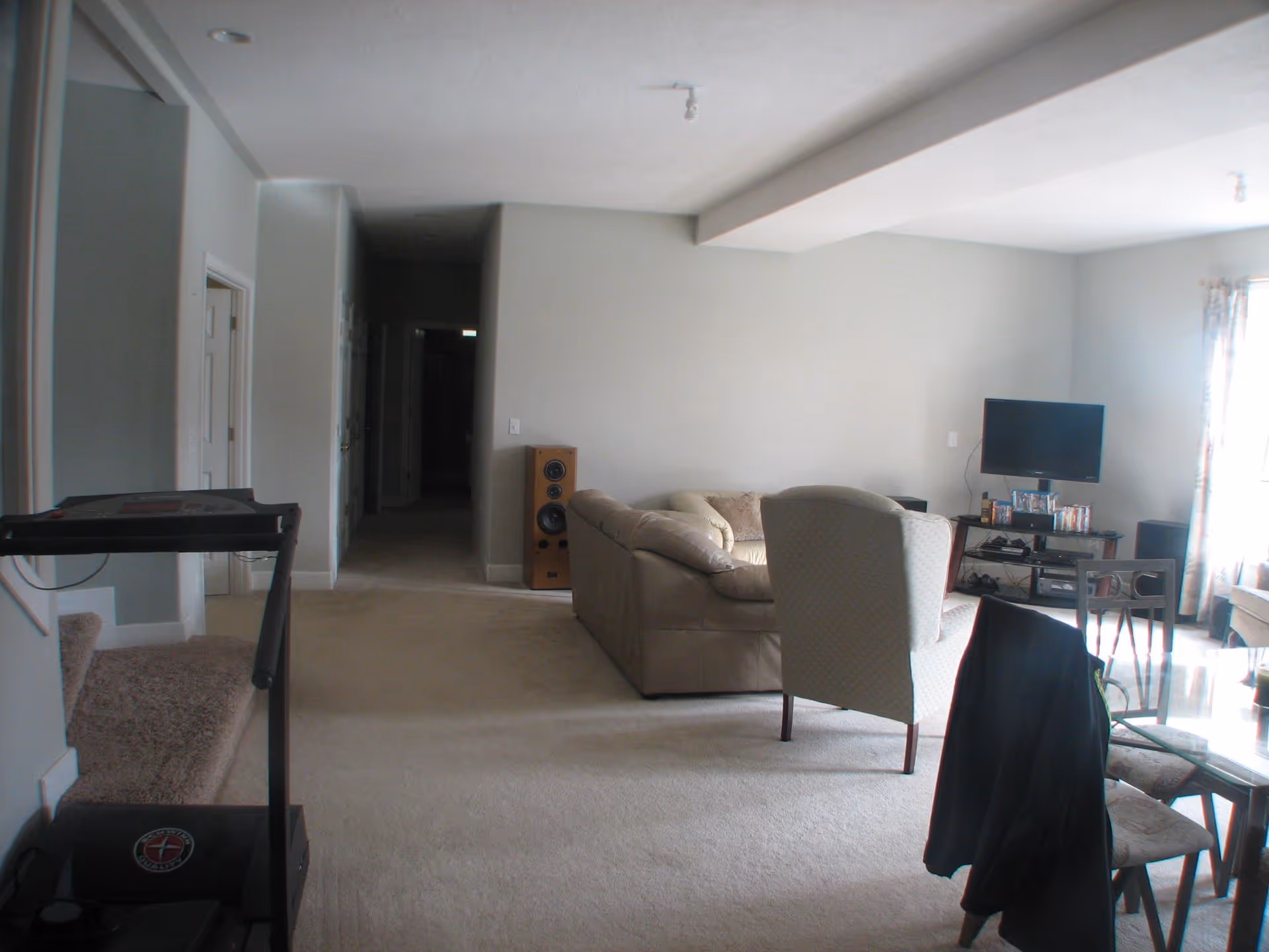Interior view of a senior living facility's common area with beige carpet, a treadmill on the left, a beige sectional sofa, a patterned armchair, a TV on a stand with DVDs, and a glass dining table with chairs. A hallway leads to other rooms in the background.