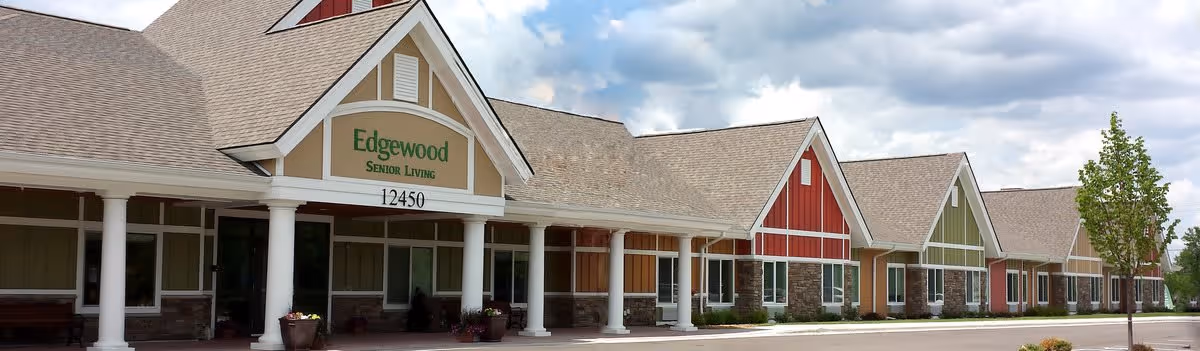 Exterior view of Edgewood Senior Living facility showing a long building with multiple peaked roofs, white columns, and a sign that reads 'Edgewood Senior Living 12450'. The building features various colored panels and large windows under a partly cloudy sky.