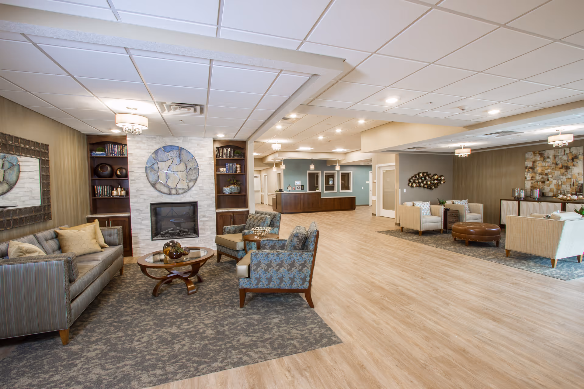A spacious and well-lit common area in a senior living facility featuring comfortable seating arrangements including sofas and armchairs around coffee tables. The room has a modern design with a fireplace, decorative wall art, built-in bookshelves, and a reception desk in the background. The flooring is a combination of wood and carpet, and the ceiling has recessed lighting and hanging light fixtures.