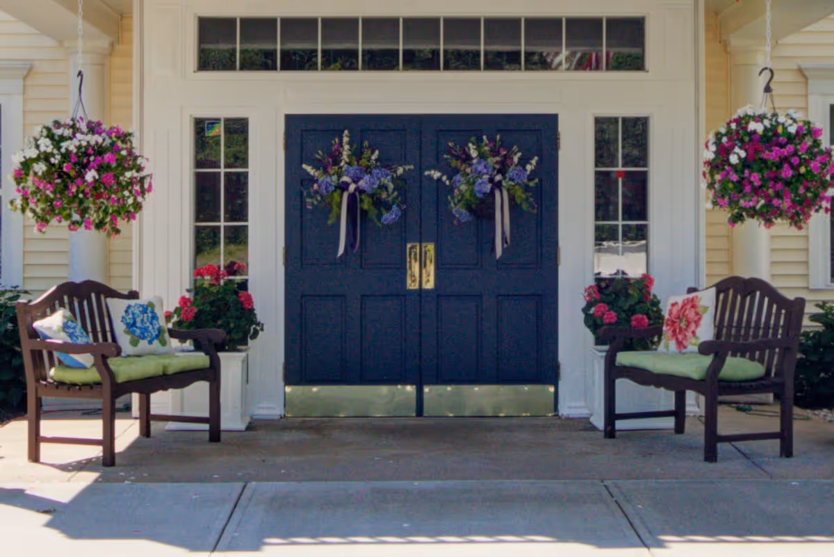 Entrance of a building with double dark blue doors decorated with floral wreaths. On each side of the doors are tall windows and white columns. Two wooden benches with green cushions and floral pillows are placed symmetrically on either side of the entrance. Hanging flower baskets with pink and white flowers are suspended above each bench.