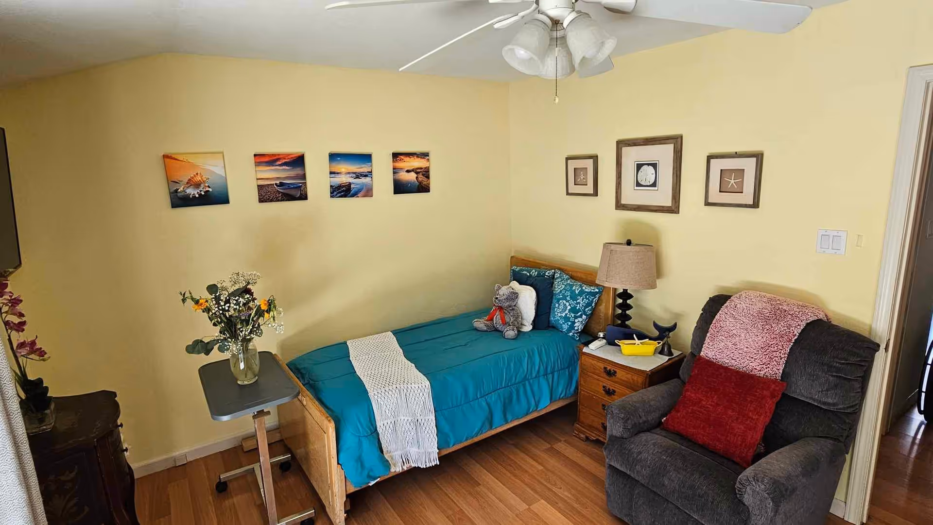 A cozy bedroom with a single bed covered in a teal blanket and a white throw. There is a teddy bear and pillows on the bed. Next to the bed is a wooden nightstand with a lamp, a small yellow boat decoration, and a whale figurine. A comfortable dark gray armchair with a red pillow and a pink throw is positioned nearby. The walls are painted light yellow and decorated with framed pictures of seashells and starfish, as well as four landscape photos above the bed. A small table with a vase of flowers is also present in the room.