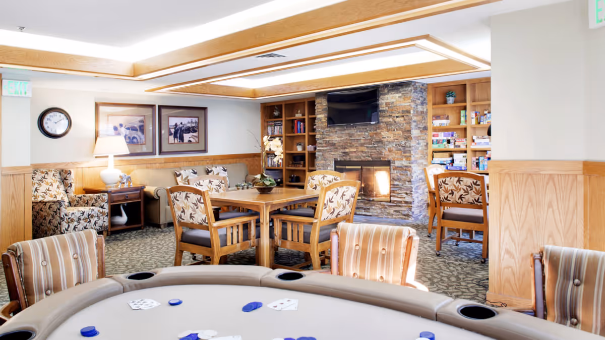Cozy senior living common room with a poker table in the foreground, wooden dining table and chairs, upholstered seating, built-in shelves and a stone fireplace.