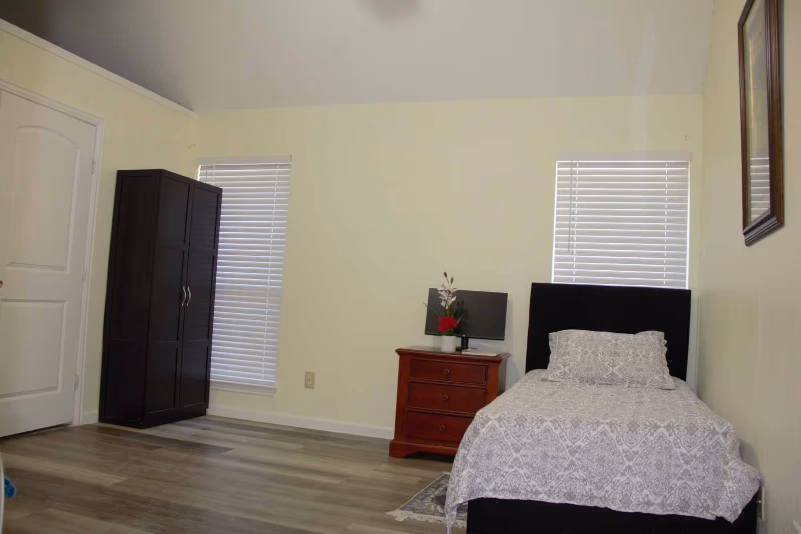 A simple bedroom with a single bed covered in a patterned gray and white bedspread, a wooden nightstand with a small monitor and a flower vase, two windows with white blinds, a dark wardrobe, and a framed picture on the wall.