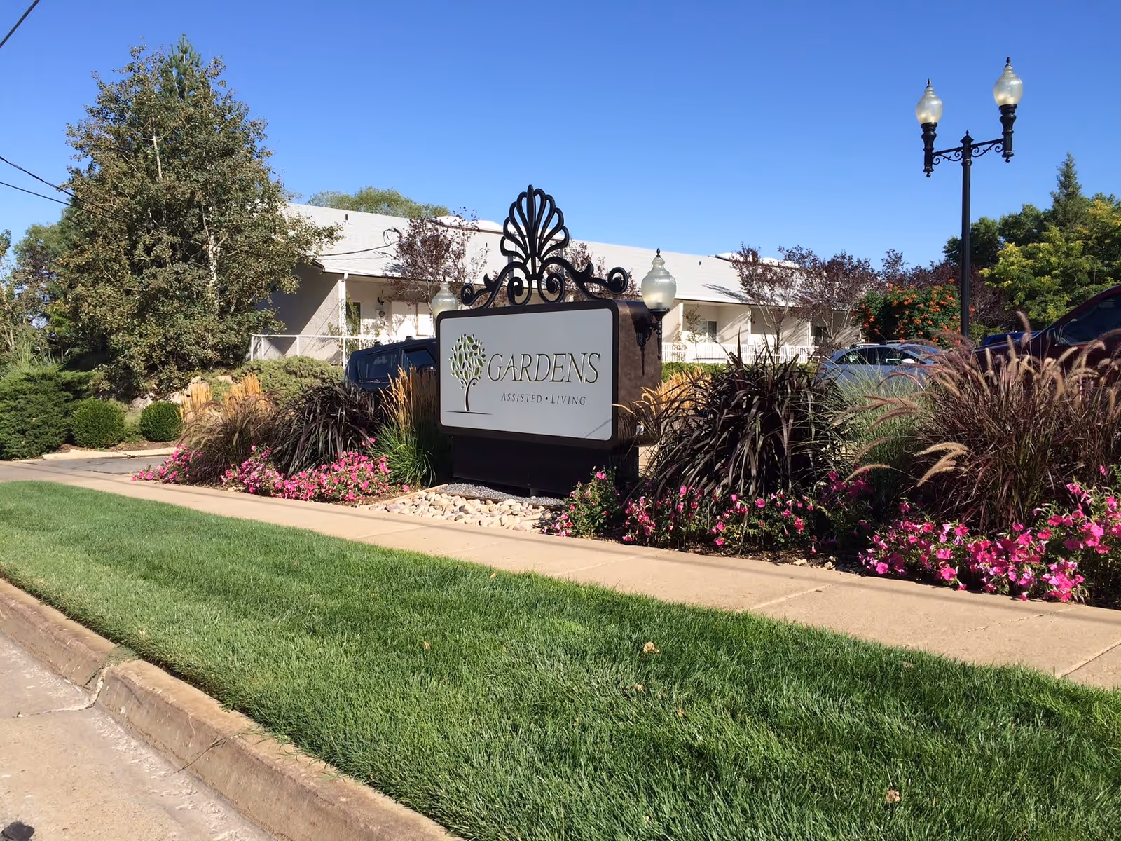 Entrance sign reading 'Gardens Assisted Living' surrounded by landscaping and flowers with the facility building in the background.