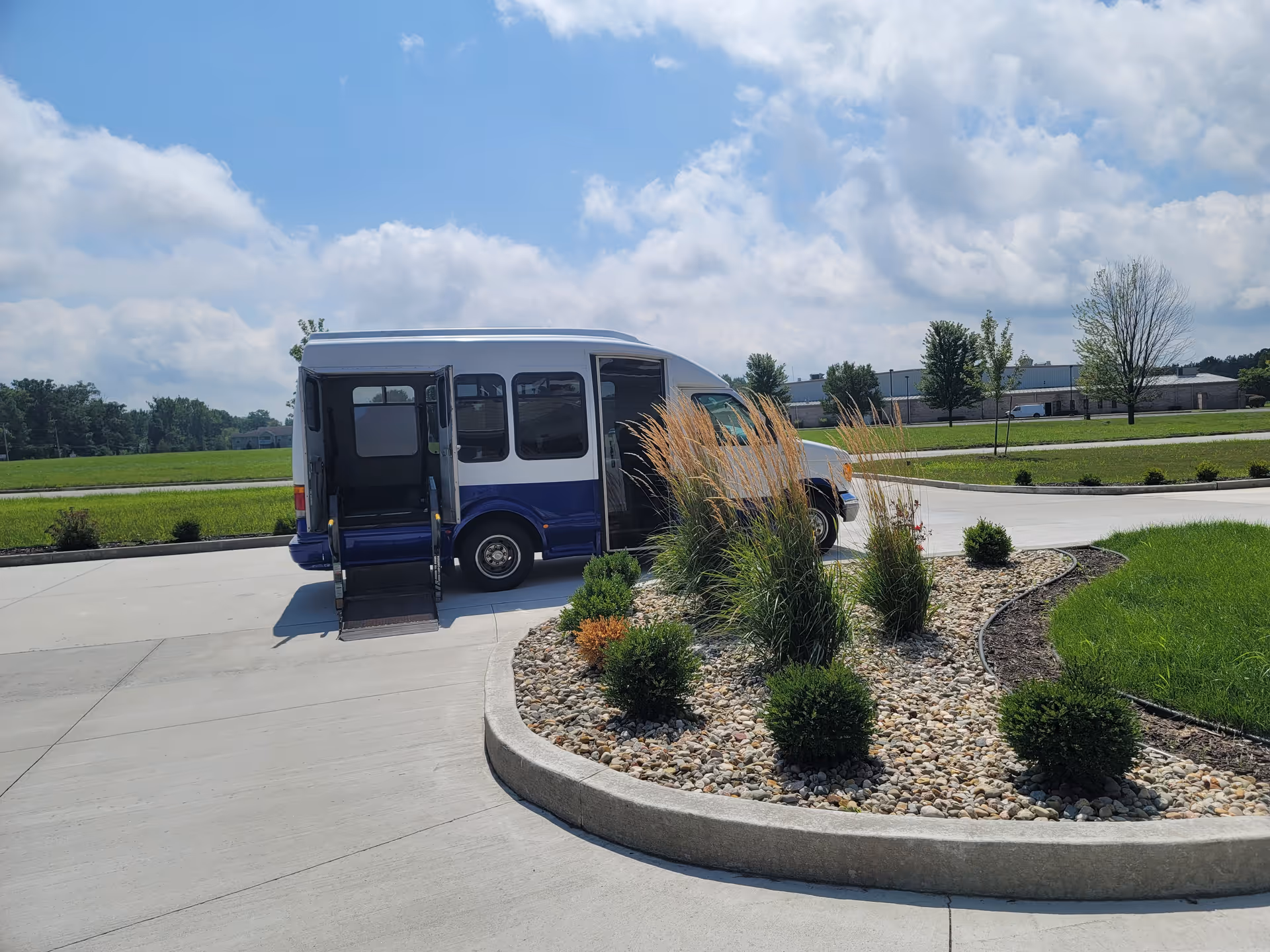 A white and blue shuttle bus with its door open and a ramp extended, parked on a curved driveway next to a landscaped area with rocks, bushes, and tall grasses under a partly cloudy sky.