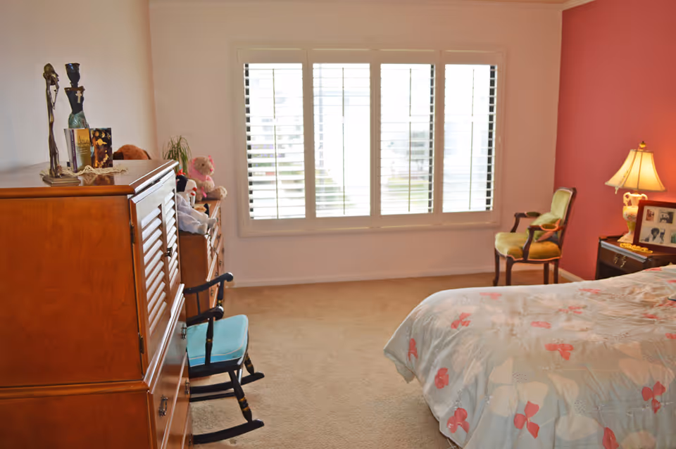 Bedroom with a large shuttered window, wooden dresser and rocking chair on the left, and a bed with a floral comforter and armchair on the right.