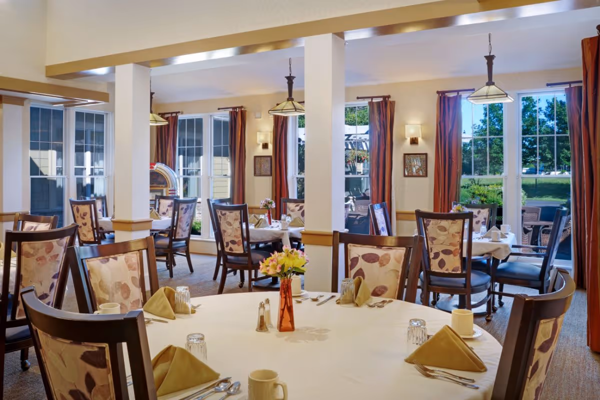 A bright dining room in a senior living facility with round tables covered in white tablecloths, set with cups, glasses, silverware, and napkins. Chairs with floral patterned upholstery surround the tables. Large windows with red curtains allow natural light to fill the room, and a vase with flowers is placed on the table in the foreground.