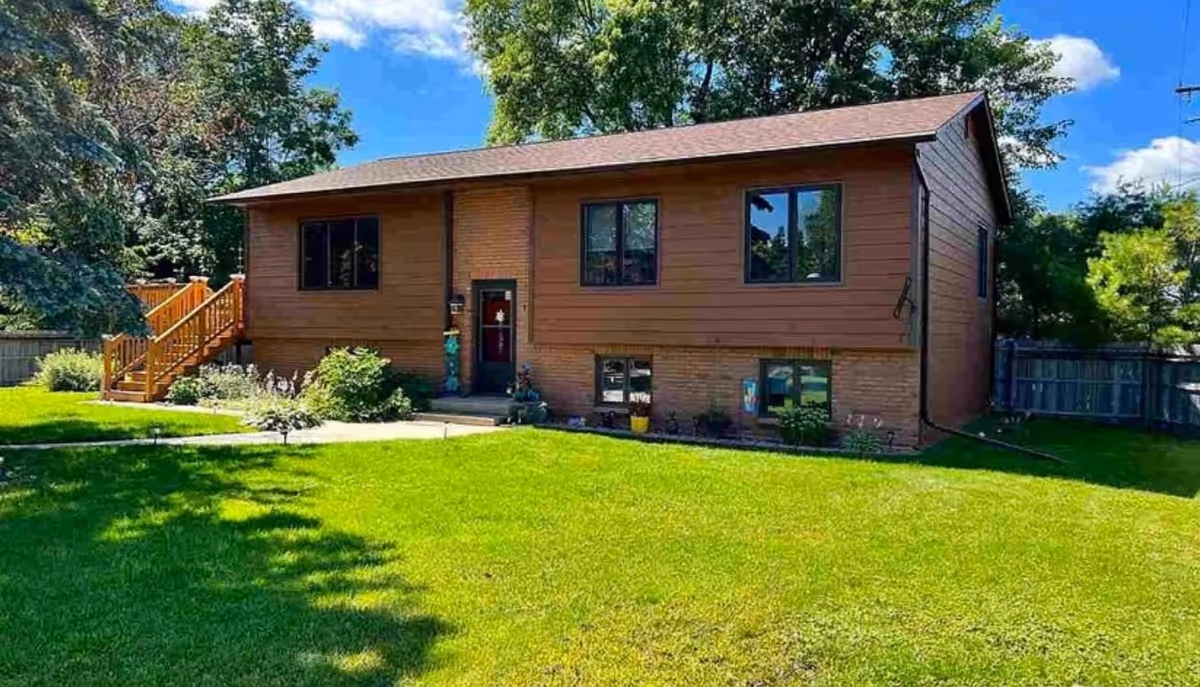 Front view of a brown split-level house with a well-kept green lawn, trees, and wooden stairs on the left.