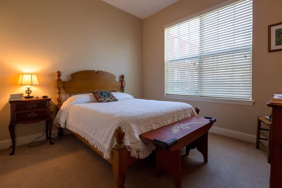 A simple bedroom with a wooden bed dressed in a white bedspread, a nightstand with a lamp, a bench at the foot of the bed, and a large window with blinds.