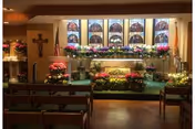 Interior of a chapel showing an altar decorated with many flowers, stained glass windows, a crucifix, and rows of pews.