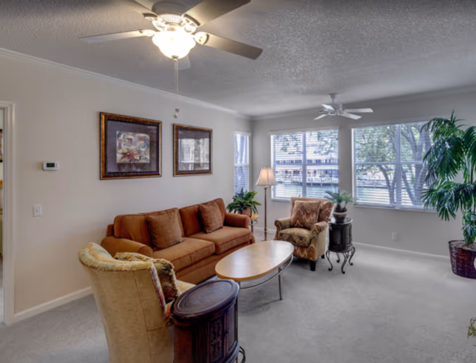 Bright living room with a sofa, multiple armchairs, coffee table, ceiling fans, and large windows overlooking trees and a building.