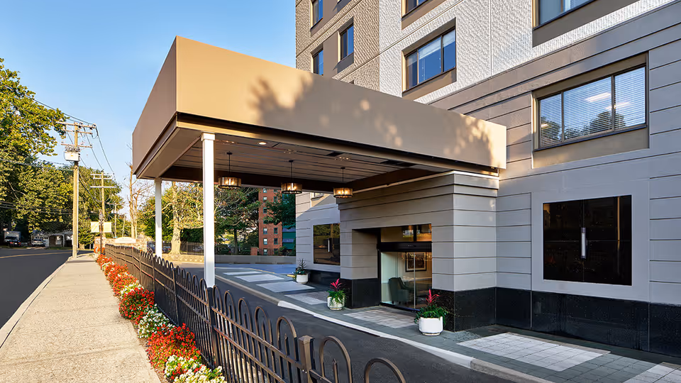 Entrance area of a multi-story building with a covered drop-off zone supported by white pillars. There are potted plants near the entrance and a black metal fence with red and white flowers along the sidewalk. Trees and power lines are visible along the street beside the building.
