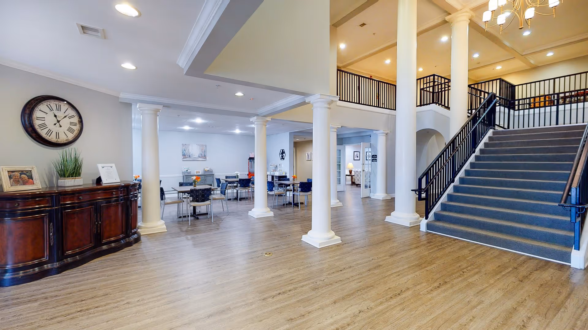 Spacious interior of a senior living facility featuring a large open area with wooden flooring, white columns, a staircase with black railings leading to an upper level, and a dining area with tables and chairs in the background. A wooden sideboard with a clock, framed photo, and plant is visible on the left wall.