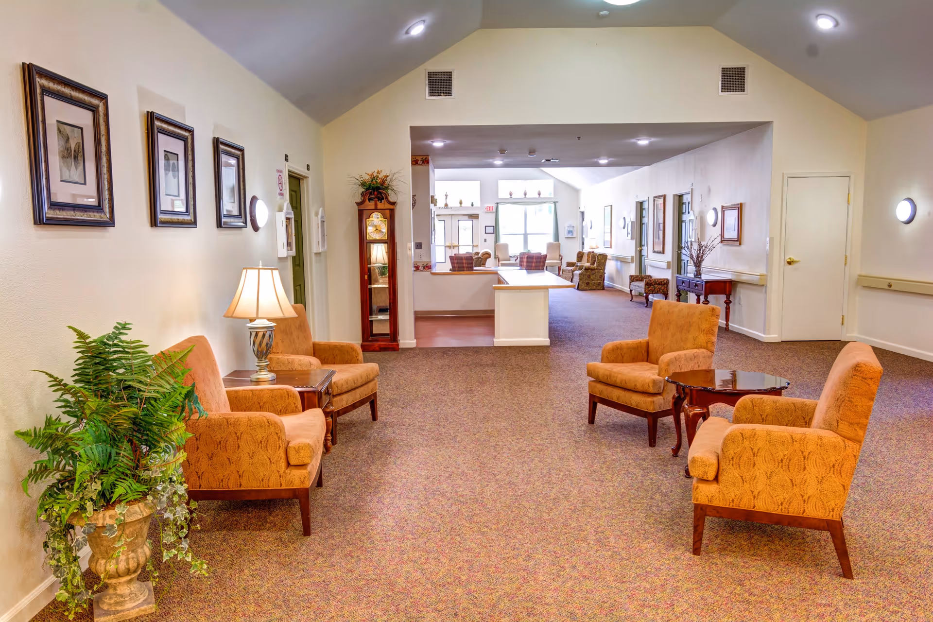 A spacious, well-lit common area in a senior living facility with beige walls and carpeted floor. The room features several orange upholstered armchairs arranged around small wooden tables. There is a tall grandfather clock against the wall, framed artwork, a table lamp, and a large potted plant. The area extends into another seating space with more chairs and large windows letting in natural light.