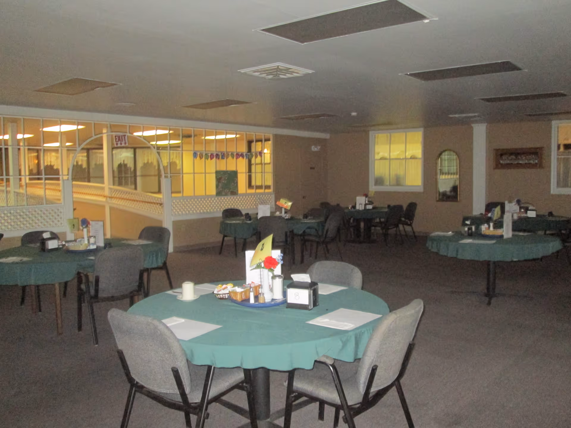 Interior view of a dining room with several round tables covered with green tablecloths, each surrounded by gray chairs. The tables have place settings, condiments, and small centerpieces. The room has beige walls, windows, and a partition with glass panels and an exit sign visible in the background.