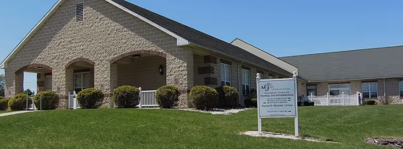 Exterior view of Emmanuel Center For Nursing, a single-story building with beige brick walls, arched entranceways, and a well-maintained green lawn. A white sign in front displays the facility's name and services.