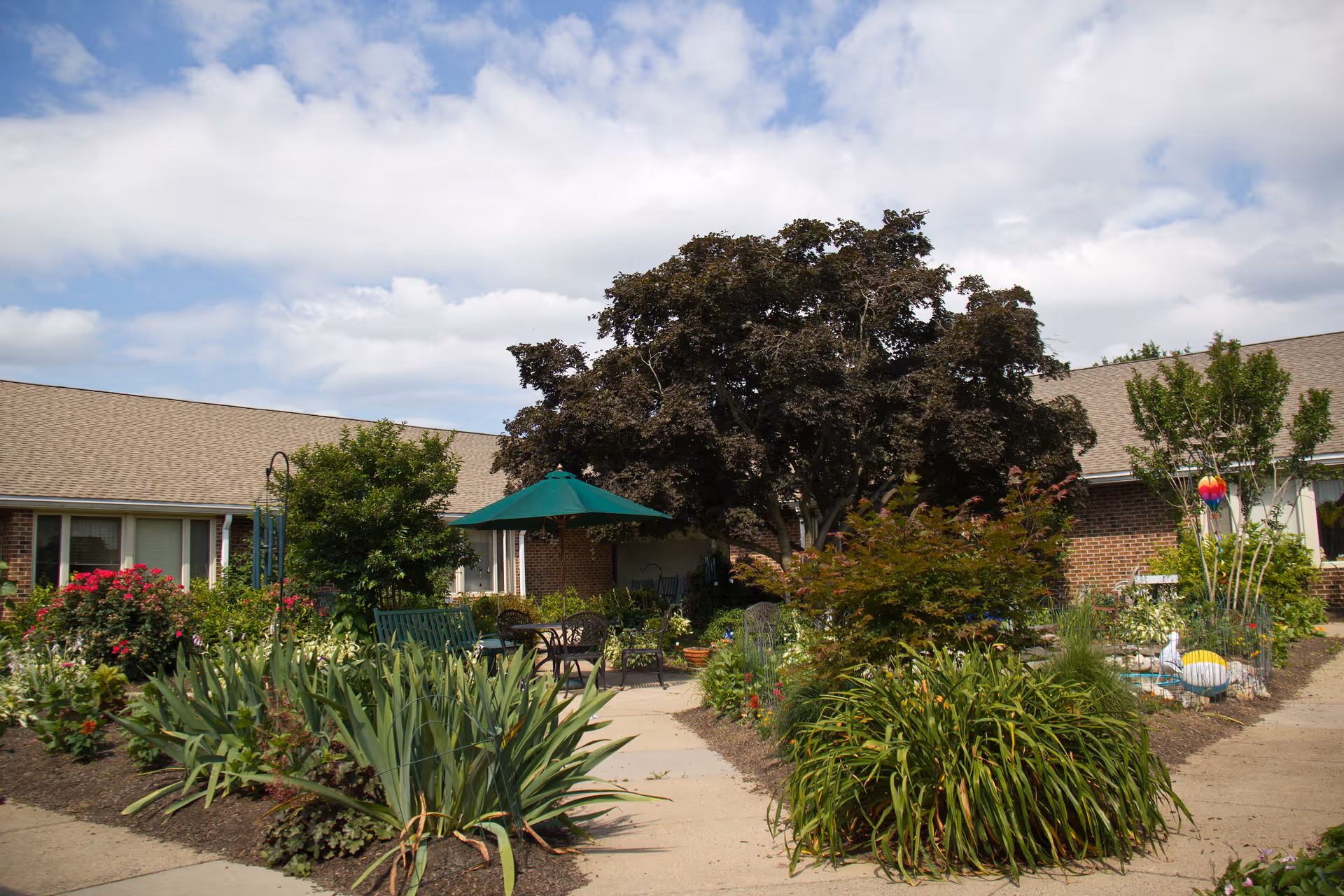 Outdoor garden area at Masonic Village at Warminster with a variety of plants, flowers, and shrubs. There is a large tree providing shade over a seating area with a green umbrella and metal chairs. The building with brick walls and windows surrounds the garden area under a partly cloudy sky.