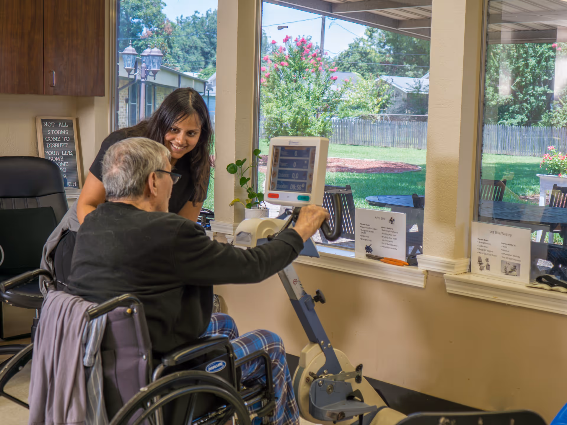 An elderly man in a wheelchair using a stationary exercise bike indoors while a woman, possibly a caregiver or therapist, smiles and encourages him. The room has large windows showing a green outdoor area with trees and flowers.