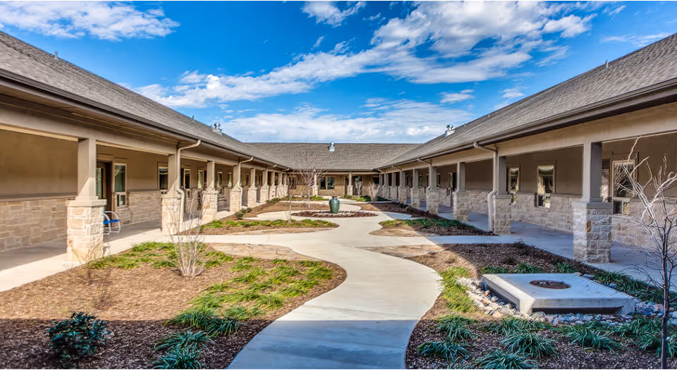 Outdoor courtyard area of a senior living facility with a winding concrete pathway, landscaped garden beds, and a fire pit. The courtyard is surrounded by single-story buildings with covered walkways and stone pillars under a partly cloudy blue sky.