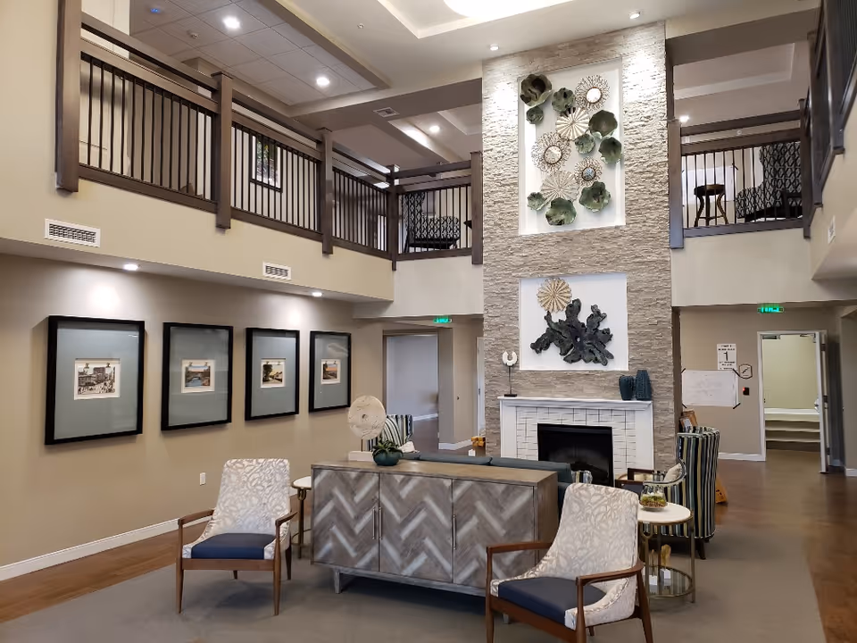 A spacious and well-lit living room area in an assisted living facility featuring a high ceiling with a second-floor balcony railing. The room has a modern fireplace with decorative art pieces above it, several framed pictures on the wall, and comfortable seating including patterned armchairs and a sofa. The floor is a combination of carpet and wood, and there are small tables with decorative items placed around the seating area.