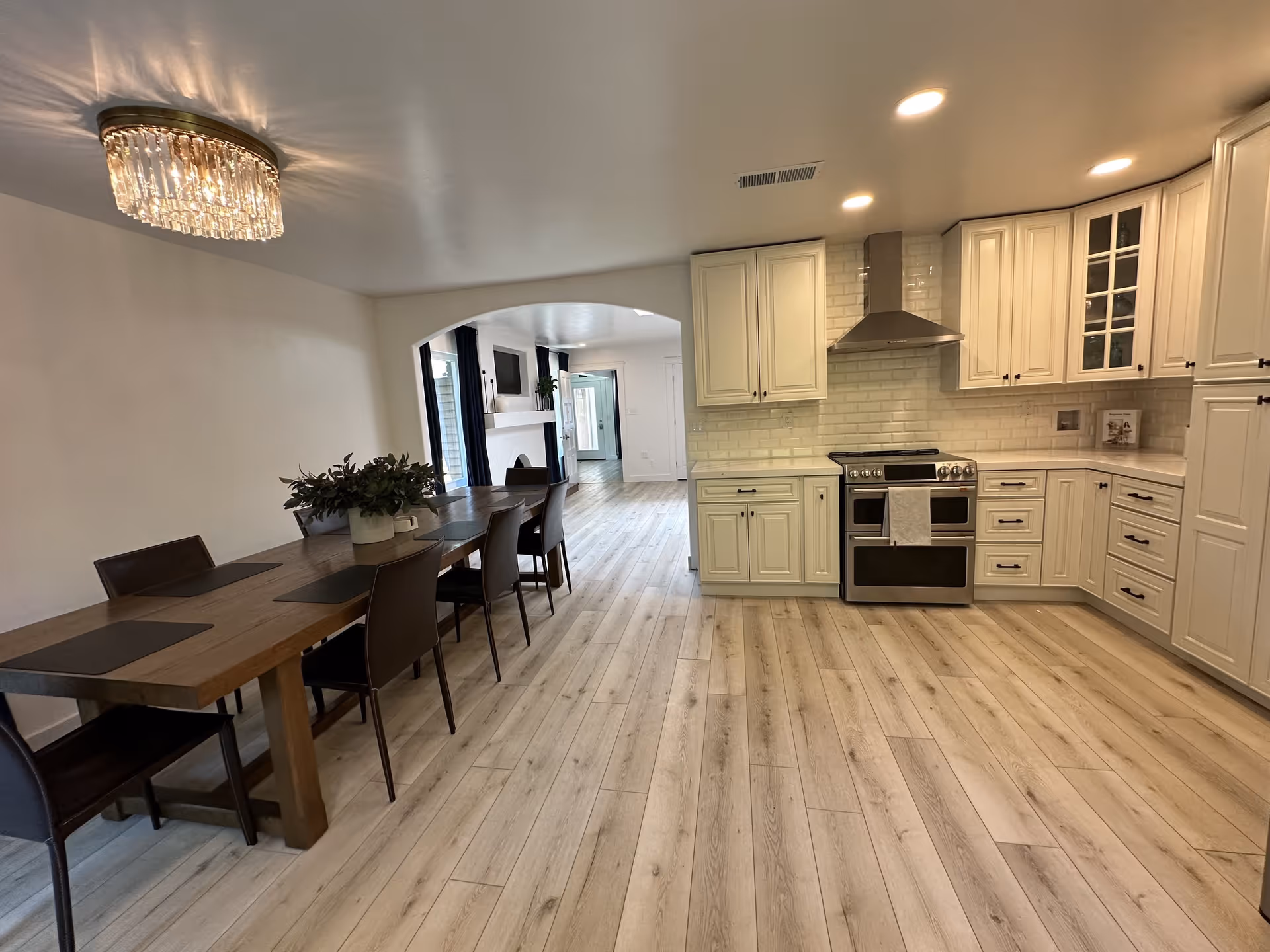A modern open-concept kitchen and dining area with light wood flooring. The kitchen features white cabinetry, a stainless steel stove with a range hood, and a white subway tile backsplash. The dining area has a long wooden table with six dark chairs and a decorative plant centerpiece. A chandelier hangs above the dining table, and an archway leads to a living room area with a fireplace and TV in the background.