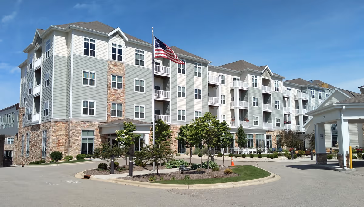 Exterior view of a multi-story senior living facility building with light green and beige siding, stone accents, multiple windows, balconies, and an American flag on a flagpole in front. The area includes landscaped greenery and a driveway under a clear blue sky.