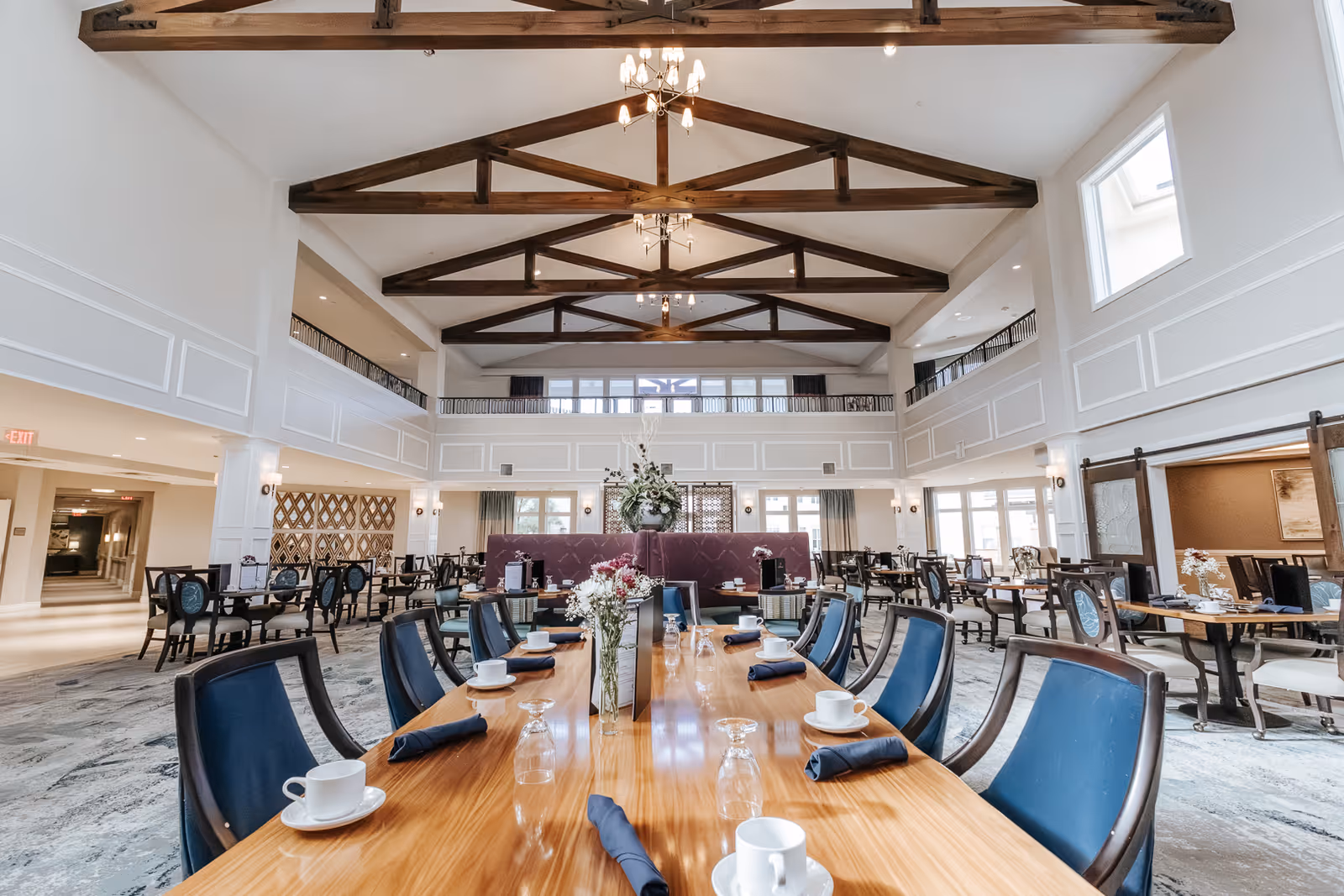 Spacious and elegant dining room with high ceilings featuring exposed wooden beams and chandeliers. The room has large windows allowing natural light to fill the space. Multiple tables are set with cups, glasses, and napkins, and a long wooden table with chairs is prominently placed in the foreground. The decor includes floral centerpieces and a mix of upholstered and wooden chairs.