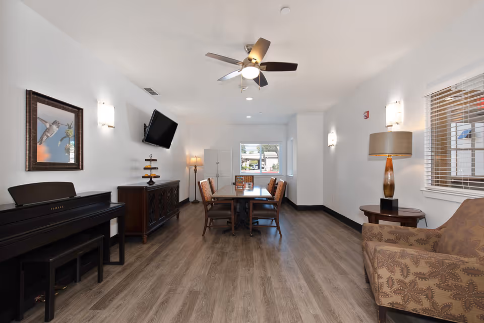 Bright communal living room with a piano on the left, a dining table and chairs in the center, a wall-mounted TV, armchair and lamp on the right, and a ceiling fan overhead.