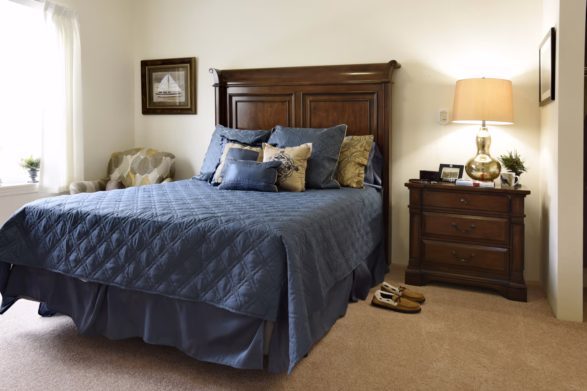 A cozy bedroom with a wooden headboard bed covered in a blue quilt and multiple decorative pillows. To the right of the bed is a wooden nightstand with a lamp, a small plant, and some personal items. A pair of slippers is placed on the carpeted floor next to the nightstand. On the left side, there is a window with white curtains and a patterned armchair beneath a framed picture of a sailboat on the wall.